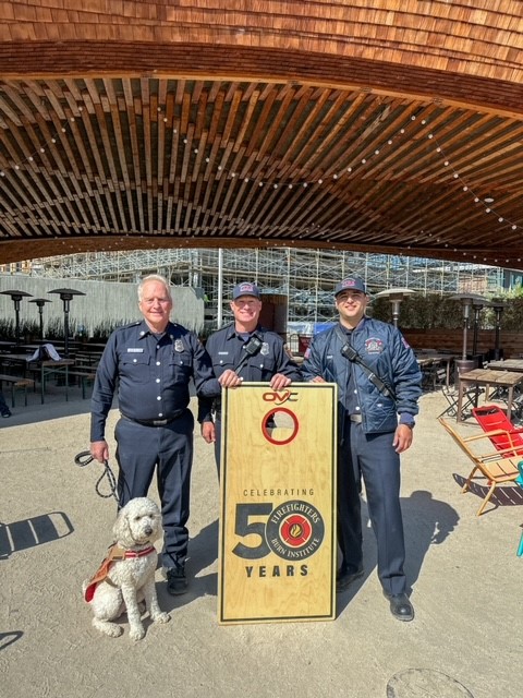 3 fireifghters standing witha cornhole board and a golden doodle promoting thee cornhole event